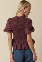 Load image into Gallery viewer, A woman shown from the back wearing a burgundy and black checkered smocked puff-sleeve top with a peplum hem, paired with blue jeans, against a solid beige background.