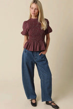 Load image into Gallery viewer, A woman modeling a short-sleeve smocked peplum top in a dark red and black textured pattern, paired with wide-leg blue jeans and black flats, against a solid beige studio background.