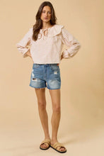 Load image into Gallery viewer, A woman modeling a light pink and white gingham long-sleeve blouse with a ruffled neckline, paired with distressed denim shorts and tan slide sandals, against a solid beige studio background.