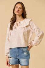 Load image into Gallery viewer, A woman modeling a light pink and white gingham long-sleeve blouse with a ruffled collar and bow tie neckline, paired with distressed denim shorts, against a solid beige background.
