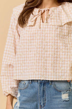 Load image into Gallery viewer, A woman wearing a pale pink and white gingham eyelet blouse with a tie-neck and ruffled collar, paired with blue jeans against a warm beige background.
