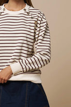Load image into Gallery viewer, A close-up of a person wearing a cream and dark brown horizontally striped long-sleeve top with decorative metallic buttons on the shoulders and upper chest, paired with dark blue denim bottoms, against a plain beige background.