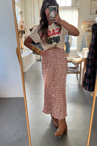 Person taking a mirror selfie wearing a graphic t-shirt and floral skirt in a room with a mirror and clothing rack.