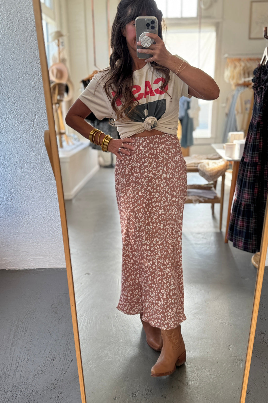 Person taking a mirror selfie wearing a graphic t-shirt and floral skirt in a room with a mirror and clothing rack.