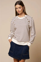 Load image into Gallery viewer, A woman modeling a cream and brown horizontally striped long-sleeve sweatshirt with decorative buttons on the shoulders, paired with a dark blue denim mini skirt, against a solid beige background.