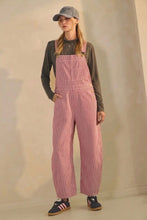 Load image into Gallery viewer, A person modeling pink corduroy bib overalls with wide legs, worn over a dark gray long-sleeve shirt, paired with a gray baseball cap and dark sneakers, standing against a plain beige wall.