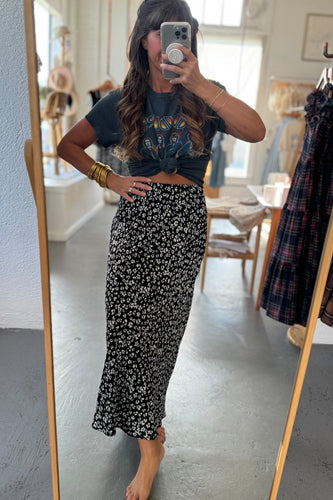 Person taking a mirror selfie wearing a dark floral skirt and crop top in a room with clothing racks.