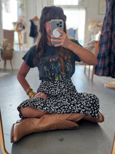 Person sitting on the floor taking a mirror selfie wearing a graphic t-shirt, floral skirt, and brown boots.