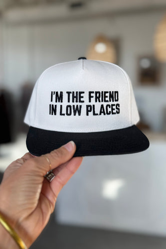Trucker hat with 'I'm the friend in low places' text held by a hand in an indoor setting.