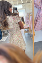 Load image into Gallery viewer, Woman trying on a floral dress in a fitting room.