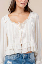 Load image into Gallery viewer, A woman wearing a cream-colored long-sleeve blouse with pleated detailing, a ruffled neckline, and a tiered hem, paired with blue jeans against a solid beige background.