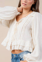 Load image into Gallery viewer, A woman wearing a cream-colored, long-sleeve ruffled blouse tucked into blue jeans, photographed against a neutral beige background.