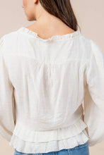 Load image into Gallery viewer, A woman viewed from the back wearing a white long-sleeve blouse with a ruffled neckline, gathered back detail, and layered ruffle hem, paired with blue jeans against a plain beige background.
