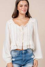 Load image into Gallery viewer, A woman wearing a cream-colored long-sleeve blouse with a ruffled neckline, tie-front detail, and gathered cuffs, paired with blue denim shorts, against a solid beige background.