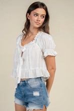 Load image into Gallery viewer, A woman modeling a white short-sleeve blouse with ruffled trim, a tie-front detail, and a ruffled hem, paired with distressed denim shorts against a plain beige studio background.