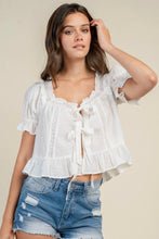 Load image into Gallery viewer, A woman wearing a white short-sleeve blouse with ruffles, lace trim, and a front tie closure, paired with distressed denim shorts, against a plain beige background.