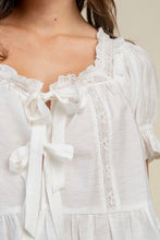 Load image into Gallery viewer, Close-up of a white textured blouse with ruffled trim, lace details, and front tie bows, worn by a model.