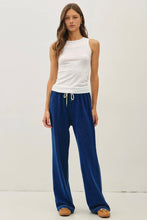 Load image into Gallery viewer, A woman modeling a pair of wide-leg blue velvet lounge pants with a white drawstring, paired with a white sleeveless top and brown shoes, against a plain white studio background.