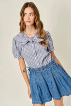 Load image into Gallery viewer, Woman wearing a checkered blouse with a bow and denim shorts on a beige background