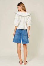 Load image into Gallery viewer, Woman wearing a white blouse with a boat neckline and blue denim shorts on a beige background