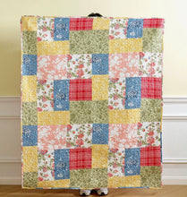 Load image into Gallery viewer, A handmade patchwork quilt held up by a person standing behind it, featuring a variety of floral, plaid, and textured square patches in blue, yellow, red, green, and pink on a cream background.