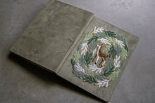 Load image into Gallery viewer, A textured sage green leather journal shown open on a gray concrete surface, featuring an intricate embroidered design of a deer within a leafy wreath on the cover.