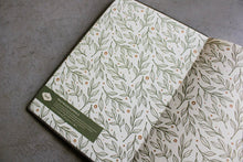 Load image into Gallery viewer, An open notebook or journal lying on a gray textured surface, showing cream-colored pages with an all-over botanical pattern of green leaves and small orange dots. A dark green label is visible on the left page.