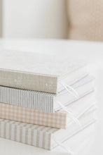 Load image into Gallery viewer, A stack of four fabric-covered books or notebooks with soft pastel patterns, including stripes, gingham, and subtle floral motifs, photographed on a light surface with a softly blurred background.