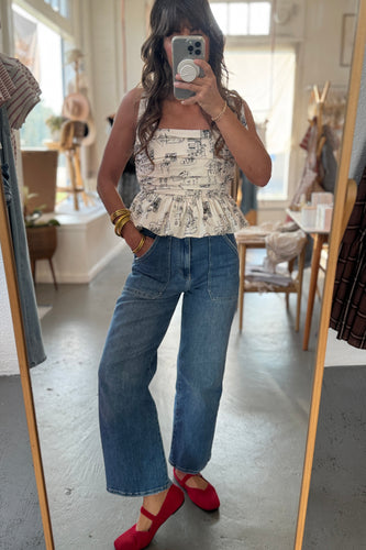 Person taking a mirror selfie wearing a patterned top, blue jeans, and red shoes in a room.