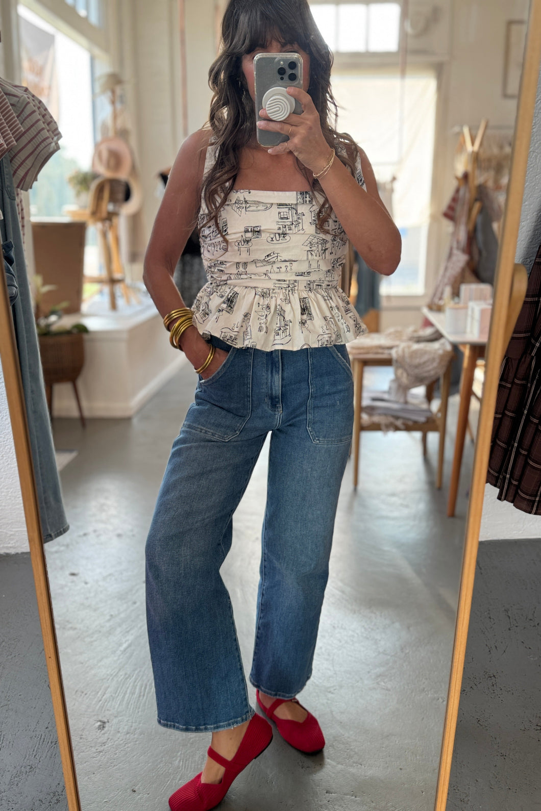 Person taking a mirror selfie wearing a patterned top, blue jeans, and red shoes in a room.