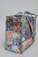 Load image into Gallery viewer, A handmade patchwork tote bag made from various colorful quilted fabric panels with floral and striped patterns, featuring blue and white striped trim and handles, shown on a neutral gray background.