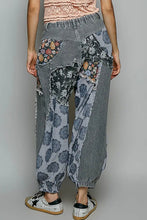 Load image into Gallery viewer, A person shown from the back wearing loose, patchwork-patterned harem-style pants with gray striped and floral fabric panels, paired with white sneakers against a plain light gray studio background.