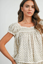 Load image into Gallery viewer, A woman wearing a cream-colored short-sleeve blouse with a small green floral print, featuring a smocked neckline and ruffled trim.
