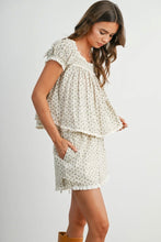 Load image into Gallery viewer, A woman wearing a matching two-piece cream and white floral print set, featuring a smocked short-sleeve top and coordinating shorts, photographed against a light gray studio background.