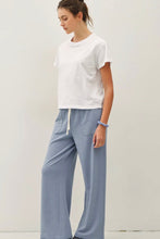 Load image into Gallery viewer, A woman wearing a white short-sleeve crewneck t-shirt and light blue wide-leg drawstring pants, standing against a plain off-white studio background.