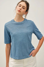 Load image into Gallery viewer, A woman wearing a short-sleeve crewneck t-shirt in a dusty slate blue color, paired with white pants, photographed against a neutral light gray background.