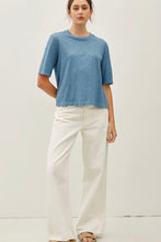 Load image into Gallery viewer, A woman modeling a short-sleeve, crew-neck blue t-shirt paired with off-white wide-leg trousers, standing against a plain white studio background.