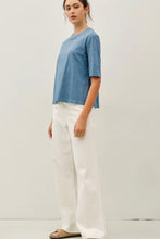 Load image into Gallery viewer, A woman modeling a short-sleeve blue t-shirt and white wide-leg pants against a plain off-white studio background.