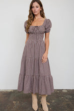 Load image into Gallery viewer, A woman modeling a brown and beige gingham midi dress with puff sleeves, a square neckline, and a tiered skirt, paired with beige ankle boots against a plain light gray wall.