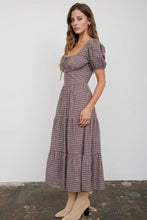 Load image into Gallery viewer, A woman modeling a brown and beige gingham midi dress with puff sleeves, a square neckline, and a tiered skirt, standing against a plain white wall.