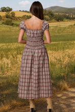 Load image into Gallery viewer, A woman stands outdoors in a rural landscape, viewed from behind, wearing a brown and beige plaid midi dress with puff sleeves and a smocked bodice.