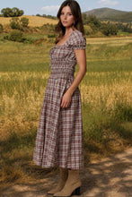 Load image into Gallery viewer, A woman stands outdoors in a grassy field wearing a brown and beige plaid midi dress with puff sleeves and a smocked bodice, paired with tan ankle boots.