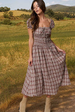 Load image into Gallery viewer, A woman wearing a brown and beige plaid midi dress with puff sleeves and a smocked bodice, standing in an outdoor grassy field.