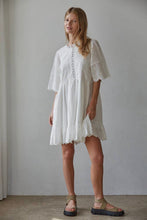 Load image into Gallery viewer, A woman modeling a white short-sleeve lace-trimmed dress with a tiered skirt, paired with brown strappy sandals, standing against a neutral light gray wall.