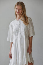 Load image into Gallery viewer, A woman wearing a white short-sleeve boho-style dress with intricate floral lace embroidery, buttoned front, and tiered ruffles, photographed against a neutral gray background.