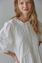 Load image into Gallery viewer, A woman wearing a white eyelet embroidered dress with puff sleeves and a buttoned front, photographed against a soft light gray studio background.