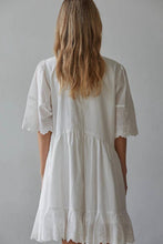 Load image into Gallery viewer, A woman viewed from behind wearing a white short-sleeve dress with eyelet lace trim on the sleeves and hem, standing against a plain light gray wall.