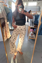 Load image into Gallery viewer, A woman takes a mirror selfie in a clothing boutique wearing beige wide-leg pants with a black bird print and a dark gray top.