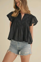Load image into Gallery viewer, Woman wearing a black blouse with ruffled sleeves and denim shorts on a beige background