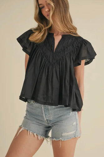 Woman wearing a black blouse with ruffled sleeves and denim shorts on a beige background